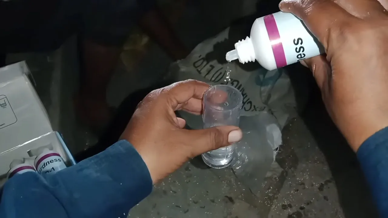 Hand squeezing drops from a dropper bottle into a small glass of water on a dark surface.